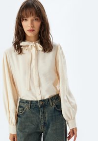 Blusa color crema con colletto a fiocco, chiusura a bottoni davanti e maniche a palloncino, abbinata a jeans in denim blu a vita alta.