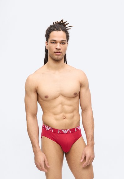 Άνδρας με γυμνό στήθος, μυώδη κορμό και κοτσίδες, φορώντας κόκκινο εσώρουχο με λευκή λωρίδα και λογότυπο στη μέση, στέκεται μπροστά σε απλό φωτεινό φόντο.