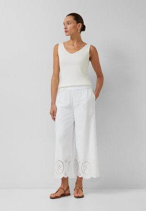 Femme portant un haut blanc sans manches, un pantalon large blanc court avec un ourlet ajouré, des sandales plates marron et des boucles d'oreilles créoles dorées, debout contre un mur gris.