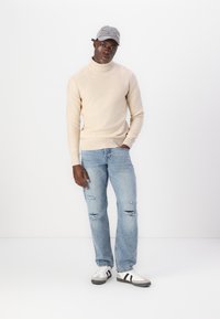Maglione beige a collo alto, aderente in vita. Jeans strappati azzurri chiari. Sneakers bianche con accenti neri. Cappello grigio con dettagli ricamati.