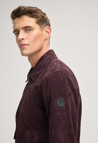 Giacca in suede bordeaux con colletto a risvolto, zipper frontale e una toppa nera rotonda sul manica sinistra. Texture liscia, design contemporaneo.