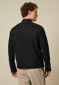 Chaqueta de ante negro con un diseño minimalista, cuello alto y puños con cremallera. Textura suave y corte entallado, vista desde la parte de atrás.