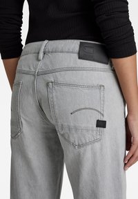 Jean en denim gris avec une texture lisse, comportant des poches arrières, un patch logo en cuir noir et des détails de couture visibles.