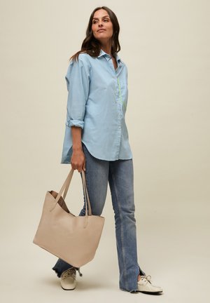 Nainen, jolla on yllään vaaleansininen nappipaita ja farkut, pitää suurta beigeä kangaskassia kädessään, seisoo yksiväristä vaaleaa taustaa vasten.