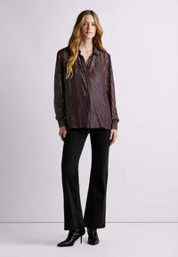 Transparante bordeauxrode blouse met verticale metallic strepen, een geknoopte kraag en lange mouwen, gecombineerd met zwarte uitlopende broek en puntige enkellaarsjes.