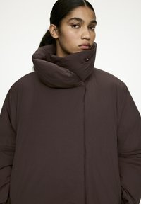 Brown puffer jacket with a high collar, featuring a button closure and a smooth texture, designed for warmth and comfort.