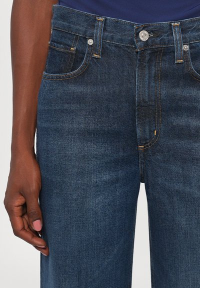 Dunkelblaue Denim-Jeans mit orangefarbenen Nähten, dargestellt an einer Person mit dunkler Haut, die ein blaues Oberteil trägt, die Hand entspannt an ihrer Seite.
