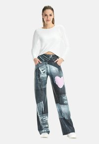 Weite Jeanshosen mit Patchwork-Design in verschiedenen Blautönen, mit ausgewaschenen Stellen und einem pinkfarbenen Herz-Akzent. Kombiniert mit einem weißen Oberteil.