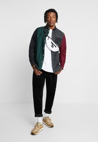Chemise à manches longues colorée en vert, noir et bordeaux, associée à un T-shirt blanc, un pantalon en velours côtelé noir et des baskets beige avec des chaussettes blanches.