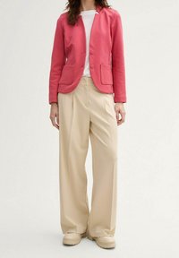Blazer en coton rose avec col cranté, deux poches avant, superposé sur un t-shirt blanc ; associé à un pantalon beige large plissé.