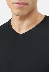 Czarna koszulka z dekoltem w serek, charakteryzująca się gładką, miękką fakturą oraz dopasowanym krojem, odsłaniająca ton skóry na dekolcie i subtelnymi detalami przeszycia.