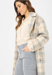 Cappotto lungo a quadri in tonalità beige e azzurro chiaro, realizzato in tessuto morbido, con colletto a bavero e due tasche frontali. Abbinato a una blusa beige e pantaloni grigi.