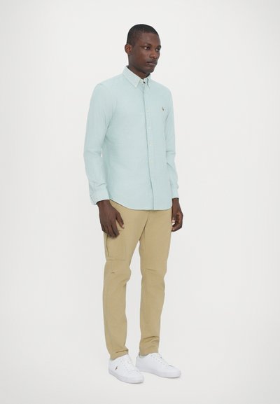 Chemise vert clair à boutons, fabriquée dans un tissu à l'apparence texturée, associée à un pantalon beige et des baskets blanches. Présente un logo sur la poitrine.