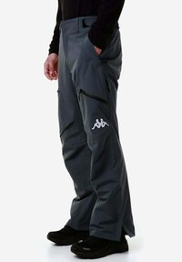 Pantaloni impermeabili grigi con vestibilità rilassata, dotati di una tasca con zip e un logo bianco sulla gamba sinistra inferiore. Indossati con calzature nere.