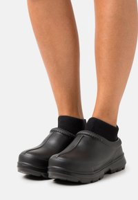 Svarta gummiklackar med en strukturerad ovandel, dämpade kanter och en robust, räfflad sula. Bärs med svarta ankelsockor som visar en avslappnad stil.