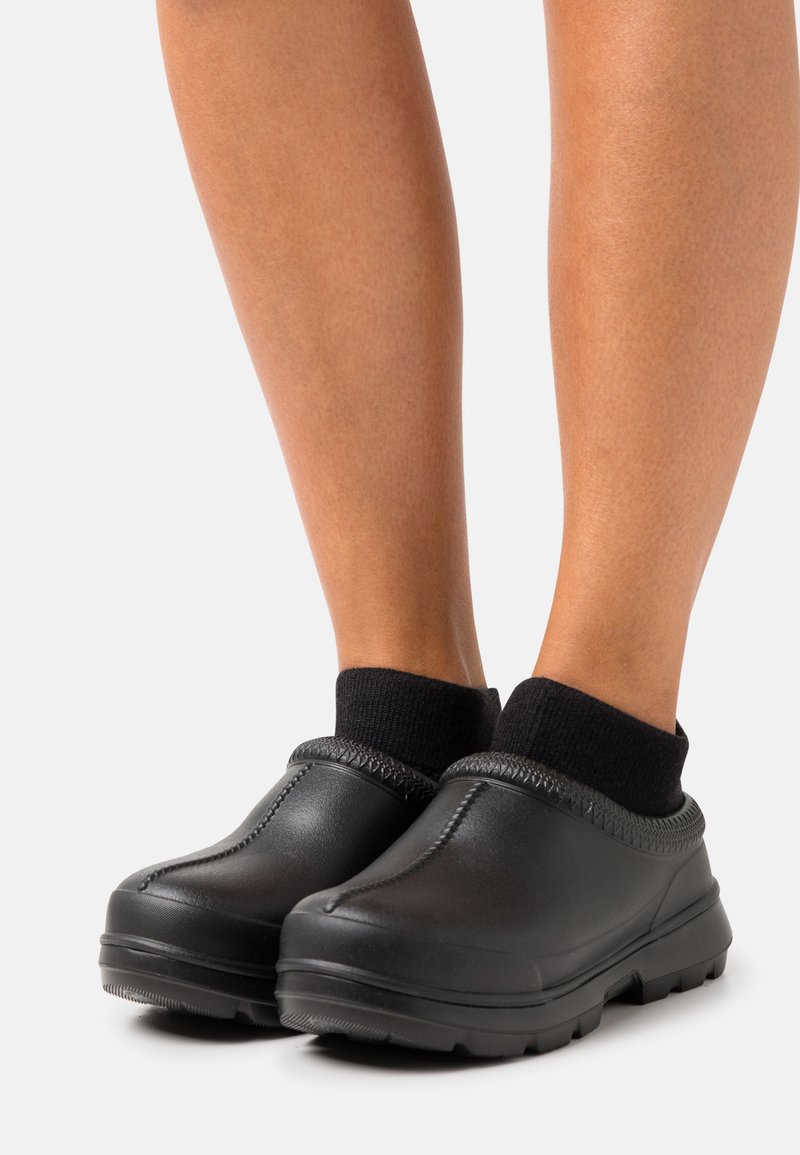 Svarta gummiklackar med en strukturerad ovandel, dämpade kanter och en robust, räfflad sula. Bärs med svarta ankelsockor som visar en avslappnad stil.