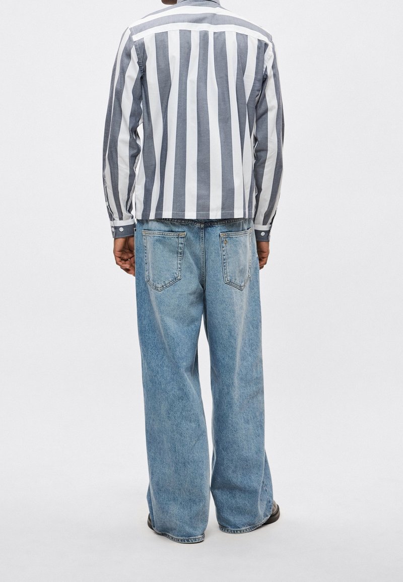 Personne portant une chemise à manches longues avec des rayures verticales blanches et grises et un jean bleu clair ample, debout dos au spectateur.