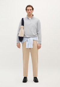 Lichtgrijze zip-up sweatshirt, beige broek, lichtblauw shirt om de taille geknoopt, zwarte schoenen, marineblauwe tas met beige accenten. Neutrale achtergrond.