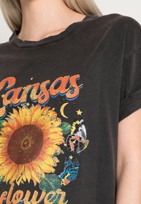 Camiseta negra de manga corta con un gráfico vibrante de girasol, coloridas mariposas y texto que dice "Girasol de Kansas". Textura de tela suave.