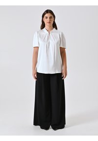 Blusa bianca a maniche corte con parte anteriore plissettata e colletto, abbinata a pantaloni neri a gamba larga. Tessuto morbido, senza motivi visibili.