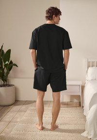 Egy férfi fekete pólóban és sportnadrágban áll, kezét zsebébe dugva, háttal egy ágy és egy cserepes növény felé, egy világos, minimalista szobában.