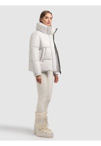 Donna che indossa una giacca imbottita grigio chiaro, leggings color crema e stivali da inverno crema con lacci, in piedi davanti a uno sfondo grigio chiaro.