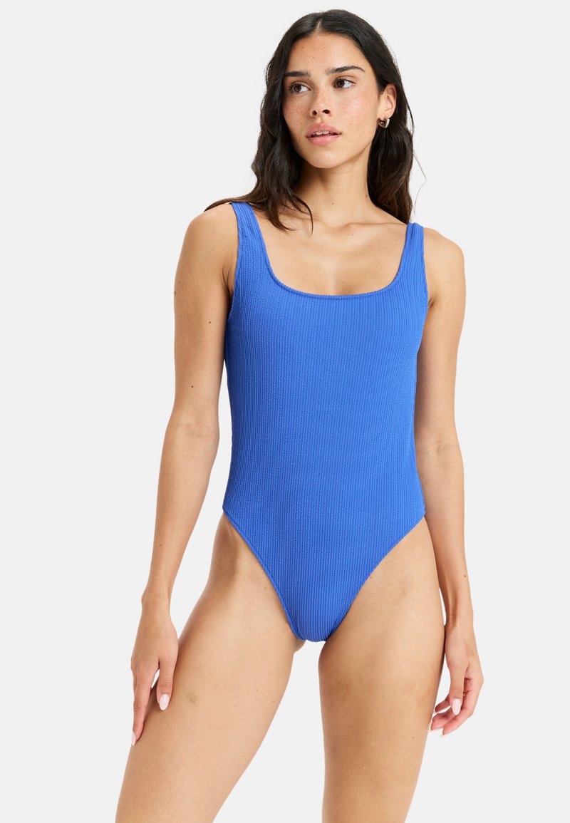 Maillot de bain une pièce bleu à côtes avec un décolleté carré et des bretelles larges. Présente des jambes haute couture et une surface texturée.