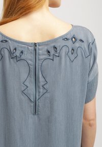 Světle modrá blůza s texturovanou tkaninou, která má zaoblený výstřih, výřezové vzory a zip vzadu.