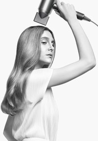 Föhn met een metalen zilveren behuizing en een grijze mondstuk boven lang, golvend haar. Het model draagt een textuur witte blouse tegen een lichte achtergrond.