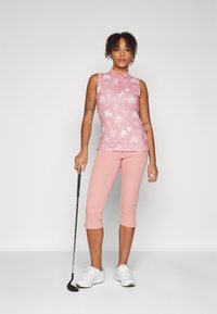 Pink sleeveless polo with a floral pattern, paired with pink capri pants. Model holds a golf club and wears white athletic shoes.