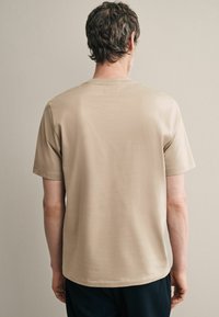Beige, rövid ujjú póló, sima textúrával és laza fazonnal, egyszerű nyakkivágással és visszafogott dizájnnal.