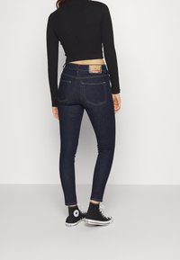 Mörkblå skinny jeans i denim, med hög midja, två bakfickor, gula sömmar och en etikett på midjebandet.