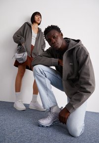 Grå zip-up hoodie med et logo, blå jeans og grå sneakers. Modellen bærer også en stribet skjorte og en brun nederdel med en grå taske.