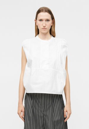 Kvinne med rett lyst brunt hår iført en hvit ermeløs bluse og svart-hvite vertikale stripete bukser, står foran en ensfarget bakgrunn.