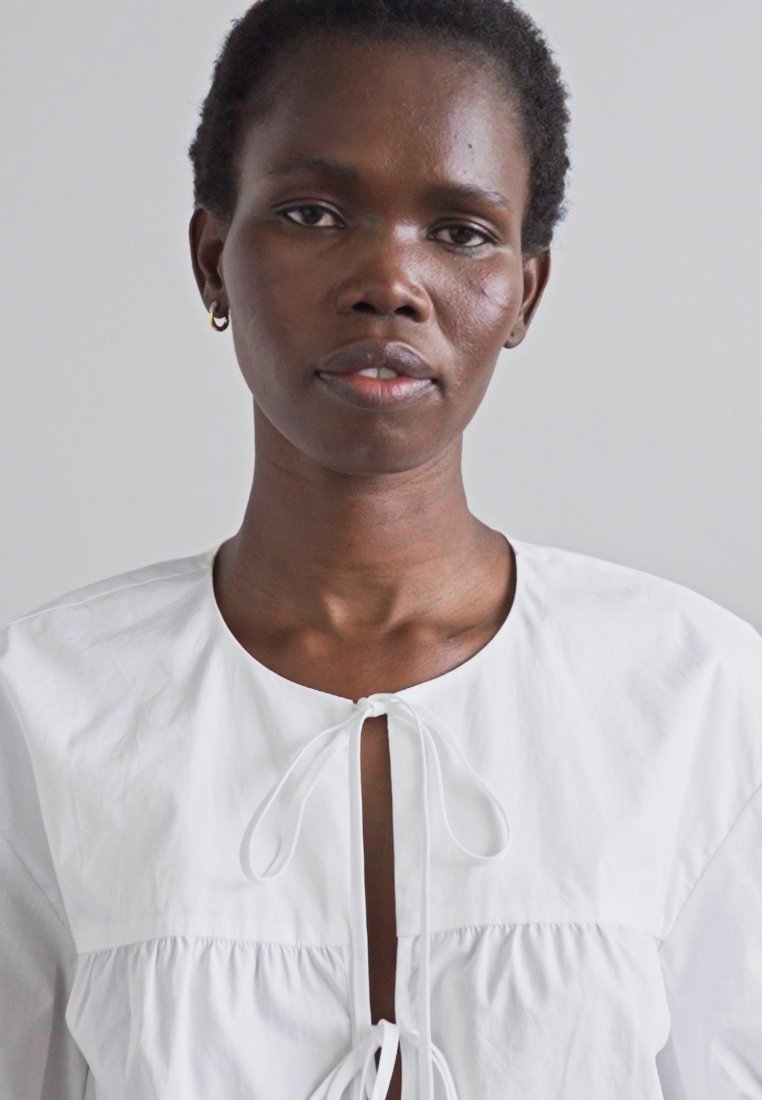 Blouse en coton blanc avec un col rond, un corsage froncé et un détail à nouer à l'avant. Texture lisse et coupe ample, les manches sont longues.