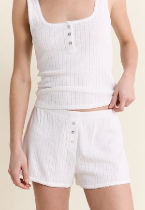 Mujer con top de punto acanalado blanco sin mangas y shorts a juego con detalles de botones, de pie contra un fondo neutral.