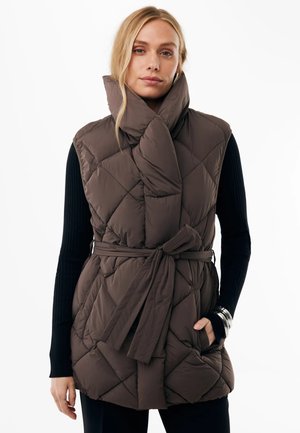 Vrouw draagt een bruine doorgestikte mouwloze bodywarmer met hoge kraag, gestrikt in de taille over een zwarte top met lange mouwen met armbanden aan één pols.