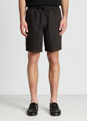 Uomo che indossa pantaloncini neri con coulisse e sandali chiusi in pelle nera, in piedi davanti a uno sfondo chiaro e uniforme.