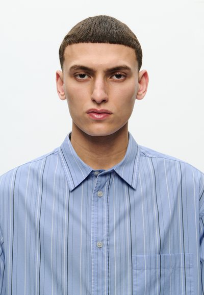 Camisa a rayas azul claro con líneas verticales negras, con cuello abotonado, bolsillo frontal y textura suave. Presenta botones metálicos.