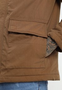 Bruine, middeldikke jas met een gladde textuur, voorzien van een grote zijzak en versterkte stiksels langs de naden.