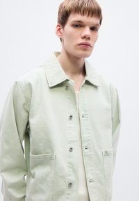 Ανοιχτοπράσινο τζιν μπουφάν με μυτερό γιακά, κούμπωμα με κουμπιά μπροστά, δύο μεγάλες τσέπες και άνετη γραμμή. Ομαλή υφή και μινιμαλιστικός σχεδιασμός.