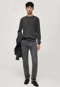 Tamno sivi vuneni džemper uparен s tamnosivim trapericama. Elegantne kožne cipele. Model drži usklađenu jaknu preko lijevog ramena. Jednostavan, moderan dizajn.