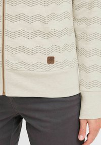 Sweatshirt zippé beige clair avec un motif zigzag texturé marron, un patch logo marron près de l'ourlet et des poignets côtelés.
