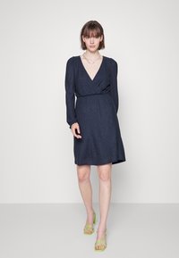 Vestido envolvente azul-marinho com mangas longas, tecido texturizado, decote em V e cintura ajustada. Usado com sapatos de salto verde com tiras.