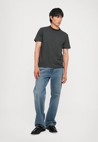 Jeune homme se tenant debout portant un t-shirt noir à motifs à manches courtes, un jean bleu clair et des chaussures en cuir noires sur un fond blanc uni.