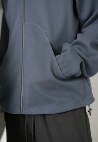 Sivá zipová bunda s rebrovým vzorom, s klokankovou vreckom, elastickými manžetami a sťahovacou šnúrkou na spodku, v kombinácii s čiernymi nohavicami.