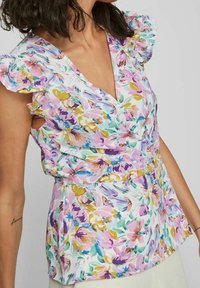 Femme portant un chemisier sans manches à fleurs avec des épaules à volants dans des tons pastel de rose, violet, vert et jaune.