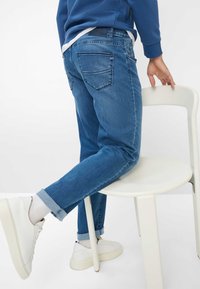 Jeans in denim blu scuro con una vestibilità slim, orli risvoltati alle caviglie e due tasche posteriori, abbinati a sneaker bianche e calzini visibili.