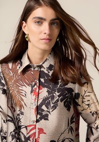 Camicia button-up con maniche lunghe di colore beige, caratterizzata da stampe botaniche in nero e rosso. Realizzata in tessuto liscio, presenta un colletto classico e bottoni bianchi.