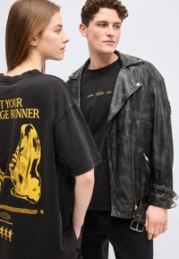 Δύο νέοι ενήλικες φορούν μαύρα graphic t-shirts· ο ένας με μαύρο δερμάτινο μπουφάν, ο άλλος με κίτρινο σχέδιο αποτυπώματος παπουτσιού στην πλάτη.